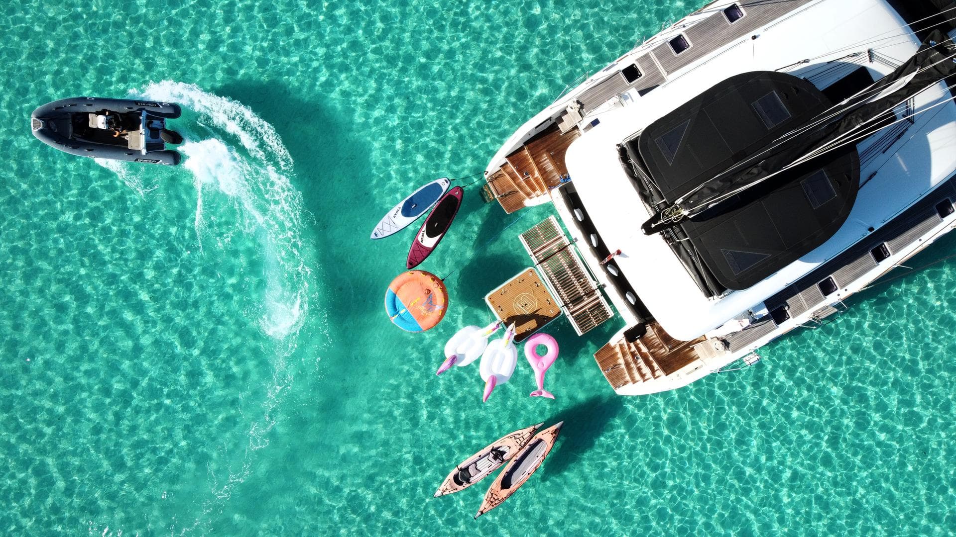 Vue aérienne d’un catamaran Lagoon au mouillage dans une eau turquoise, entouré de paddles, kayaks et jouets nautiques